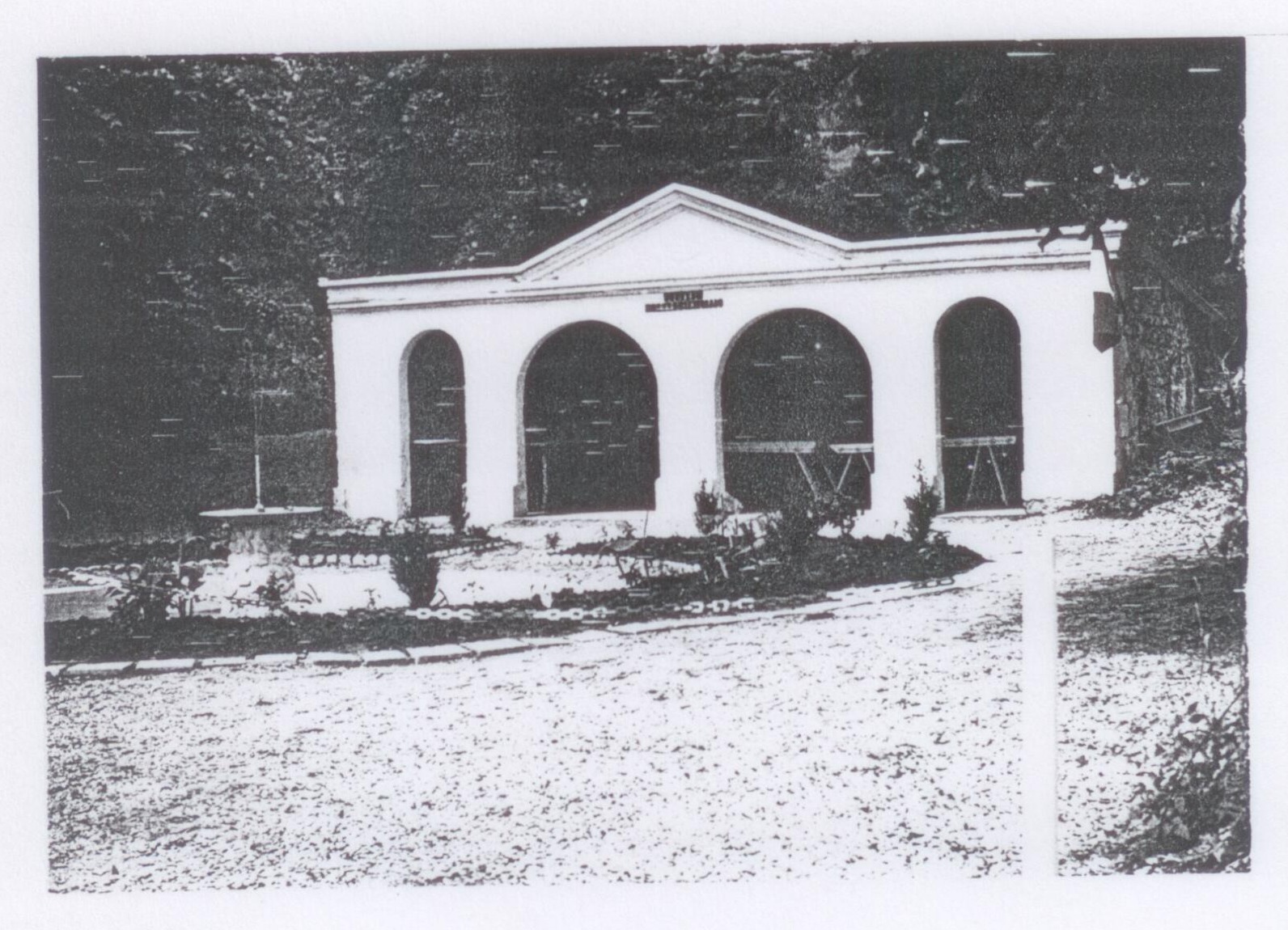 Photographie de l'ancien lavoir de Merlieux devenu square Gérard-Philipe : façade claire à quatre arcades sous un fronton, devant une allée et des massifs